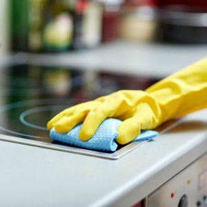 people, housework and housekeeping concept - close up of woman hand in protective glove with rag cleaning cooker at home kitchen